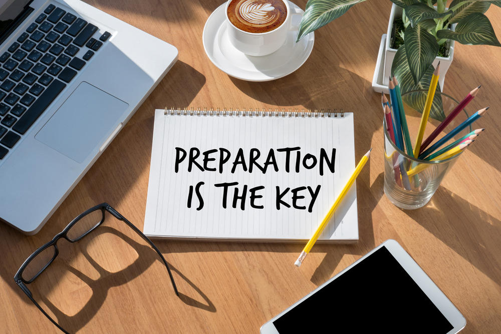 Notebook on a desk with "preparation is the key" written in bold black ink. A laptop, pencils, tablet and eye glasses are nearby.