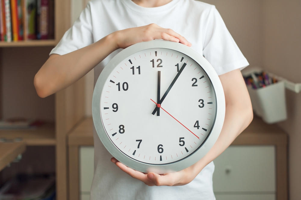 A teenager boy is holding a big clock with big numbers. A child in a white T-shirt, a watch close-up, a person is not visible. Concept mode, schedule, rest.