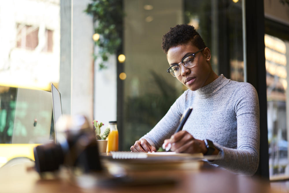 Young professional African American woman wearing a sweater, sitting at a table in a coffee shop writing in a notebook.