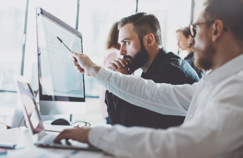 Two men in business attire looking at a computer monitor, one pointing with a pen.
