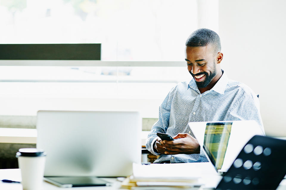 Man looking at cell phone in front of a computer while grinning.