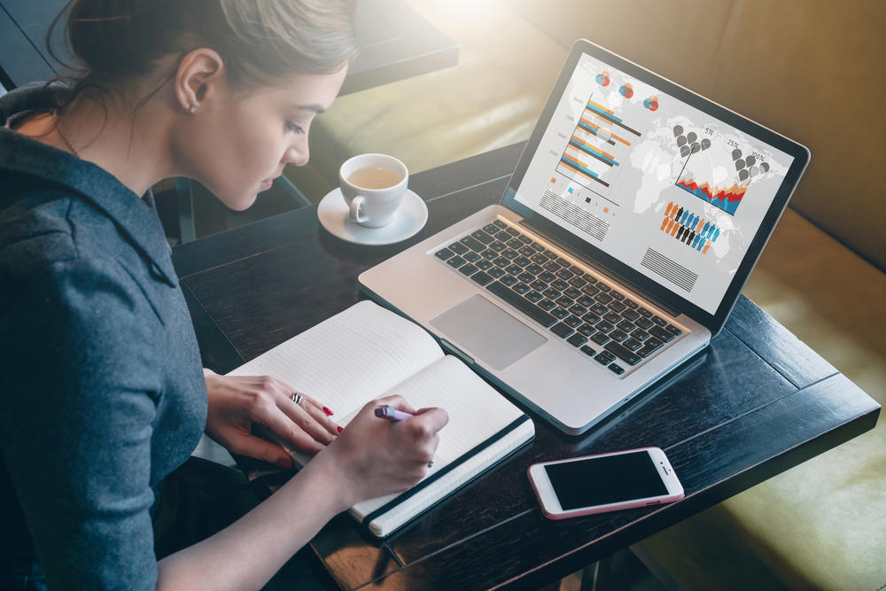 Young business woman sitting at table and taking notes in notebook. On table is laptop, smartphone and cup of coffee. On computer screen graphics and charts.