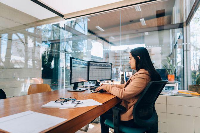 woman with dark hair sitting at a desk with a spreadsheet on two monitors and she is using copilot ai in accounting