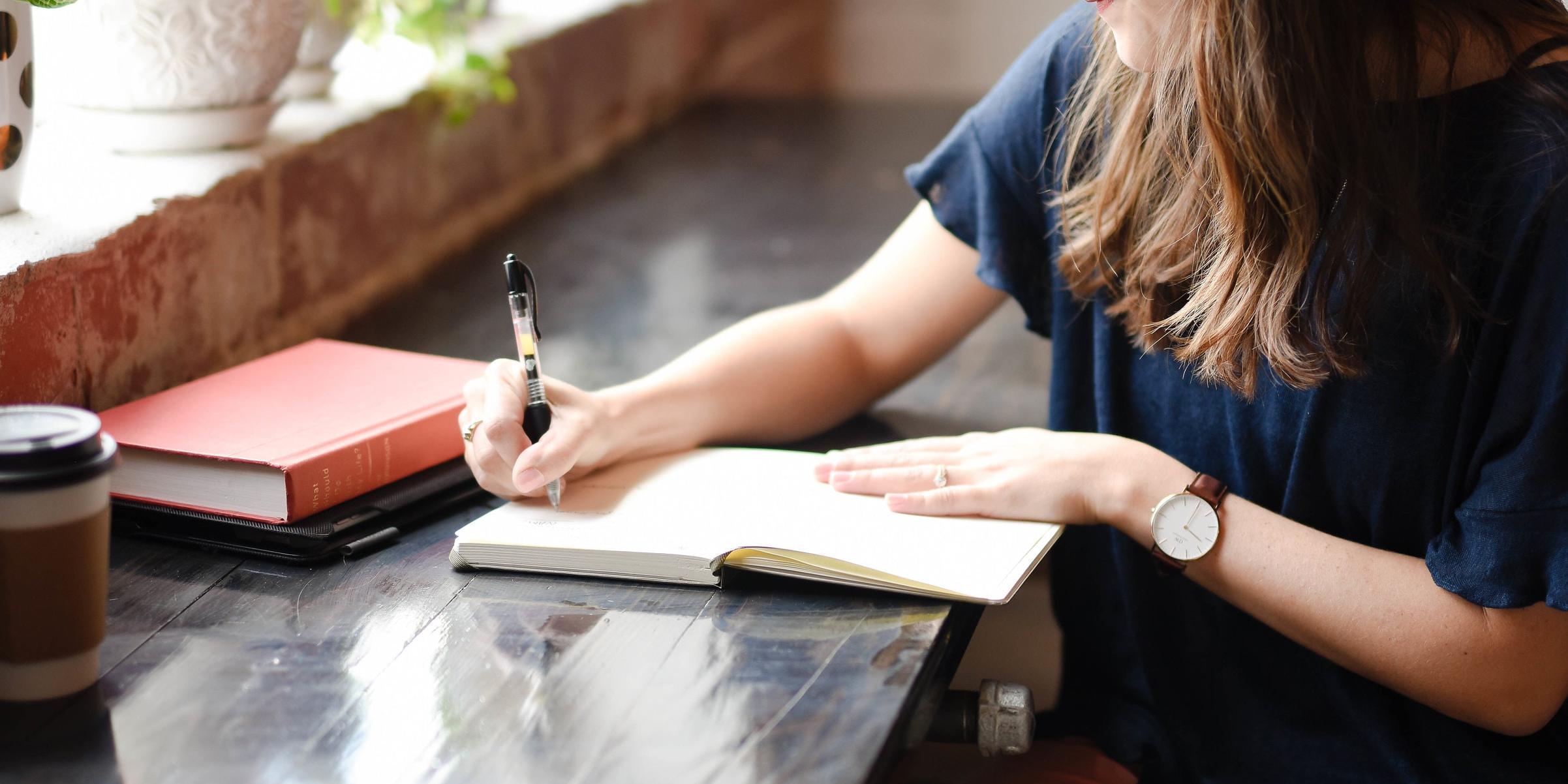 Young woman sits at desk under sunny window ledge writing in a journal.